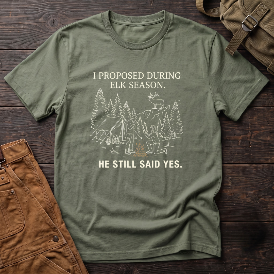 Military Green funny elk hunting t-shirt with I Proposed During Elk Season, He Still Said Yes text and a line-art hunter kneeling by the campfire at an elk camp tent, with a bull elk on the ridge