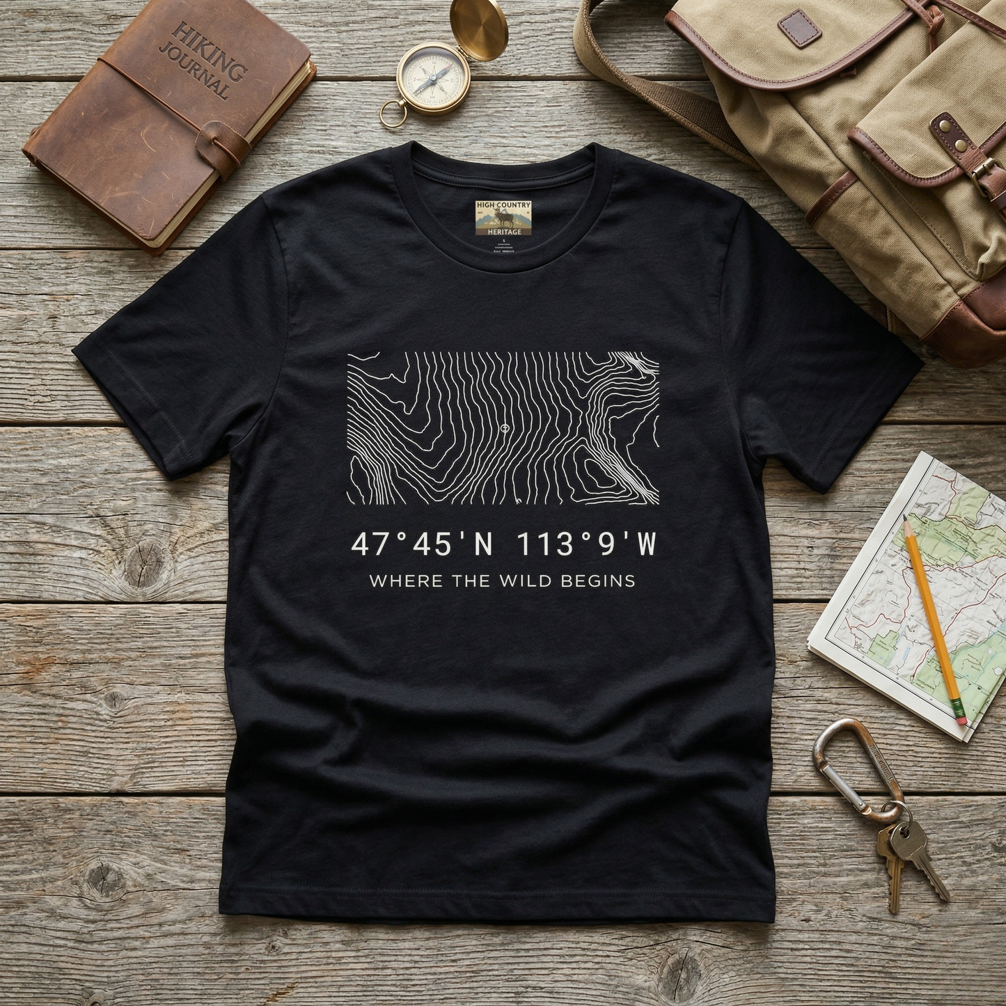 Flat lay of a black Bob Marshall Wilderness coordinates t-shirt on a weathered wood surface alongside a hiking journal, brass compass, canvas pack, and topo map, showing the minimalist contour line design and coordinates 47 degrees 45 N 113 degrees 9 W.