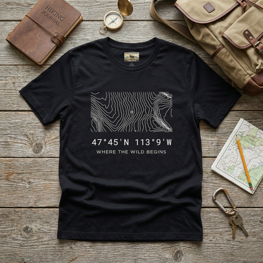Flat lay of a black Bob Marshall Wilderness coordinates t-shirt on a weathered wood surface alongside a hiking journal, brass compass, canvas pack, and topo map, showing the minimalist contour line design and coordinates 47 degrees 45 N 113 degrees 9 W.