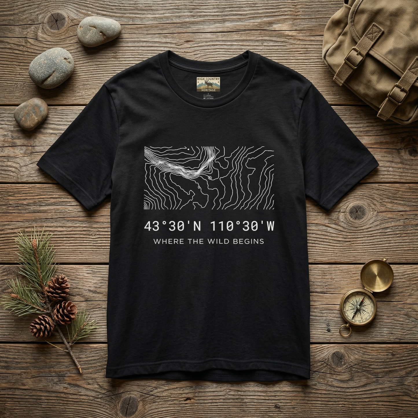 Flat lay of a black Bridger-Teton elk hunting coordinates t-shirt on a weathered wood surface with river stones, pine cones, a brass compass, and a canvas pack, showing the minimalist contour map design and coordinates 43 degrees 30 N 110 degrees 30 W.