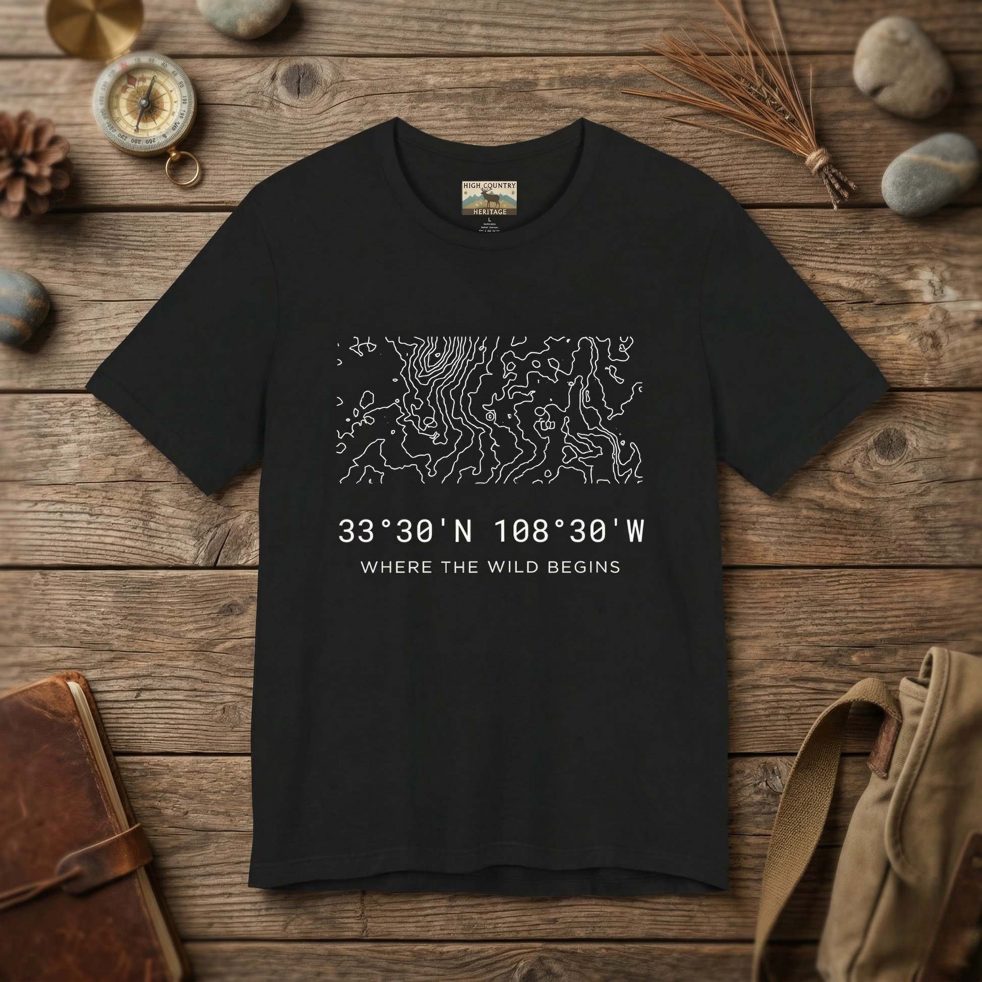 Flat lay of a black Gila Wilderness elk hunting coordinates t-shirt on a weathered wood surface alongside a brass compass, leather journal, pine cone, and canvas pack, showing the complex minimalist contour map design and coordinates 33 degrees 30 N 108 degrees 30 W.