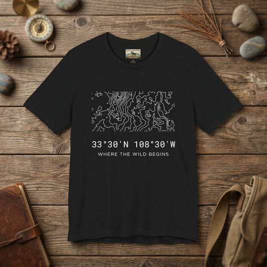 Flat lay of a black Gila Wilderness elk hunting coordinates t-shirt on a weathered wood surface alongside a brass compass, leather journal, pine cone, and canvas pack, showing the complex minimalist contour map design and coordinates 33 degrees 30 N 108 degrees 30 W.
