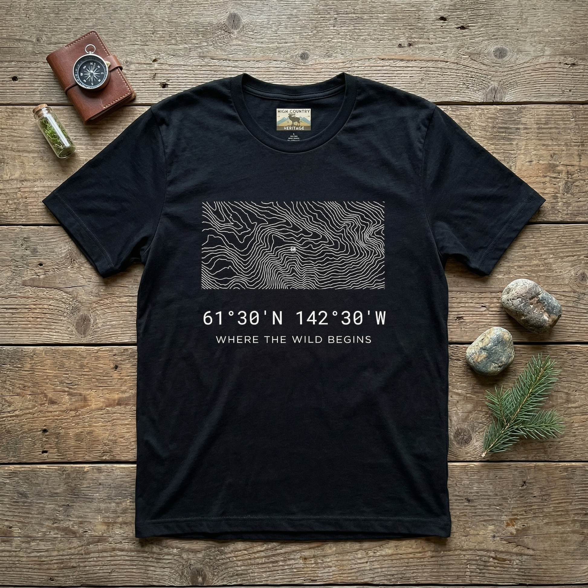 Flat lay of a black High Country Heritage Alaska Wrangell-St. Elias topographic map t-shirt on a weathered wood surface alongside a leather compass wallet, small glass vial, and river stones, showing the minimalist flowing contour map design and coordinates 61 degrees 30 N 142 degrees 30 W.