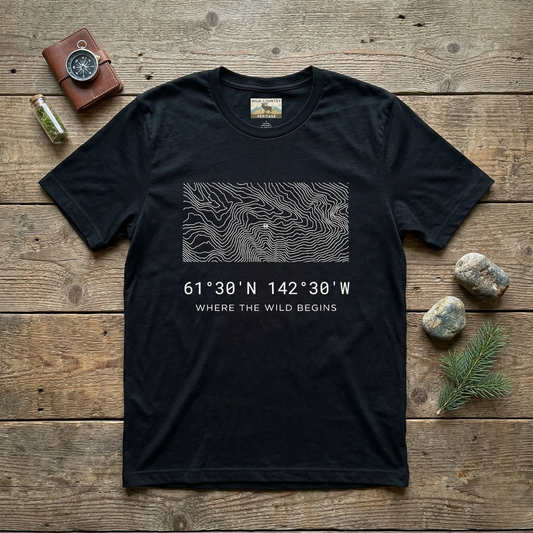 Flat lay of a black High Country Heritage Alaska Wrangell-St. Elias topographic map t-shirt on a weathered wood surface alongside a leather compass wallet, small glass vial, and river stones, showing the minimalist flowing contour map design and coordinates 61 degrees 30 N 142 degrees 30 W.