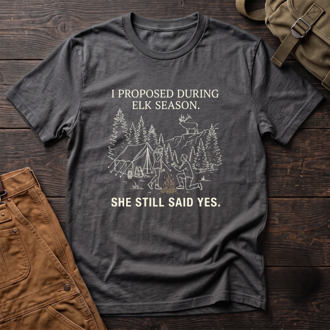Asphalt funny elk hunting t-shirt with I Proposed During Elk Season, She Still Said Yes text and a line-art hunter kneeling by the campfire at an elk camp tent, with a bull elk on the ridge