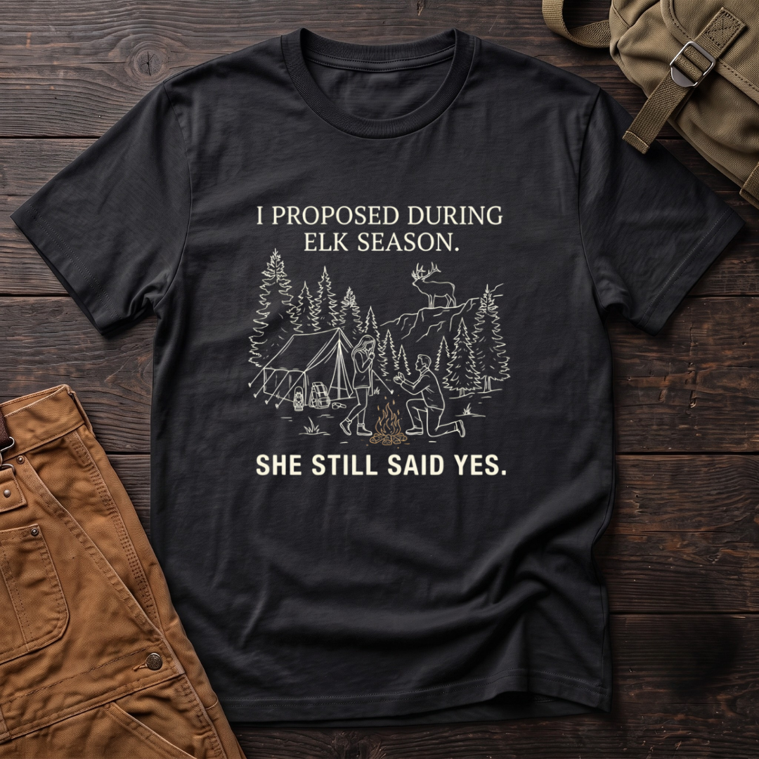 Black funny elk hunting t-shirt with I Proposed During Elk Season, She Still Said Yes text and a line-art hunter kneeling by the campfire at an elk camp tent, with a bull elk on the ridge