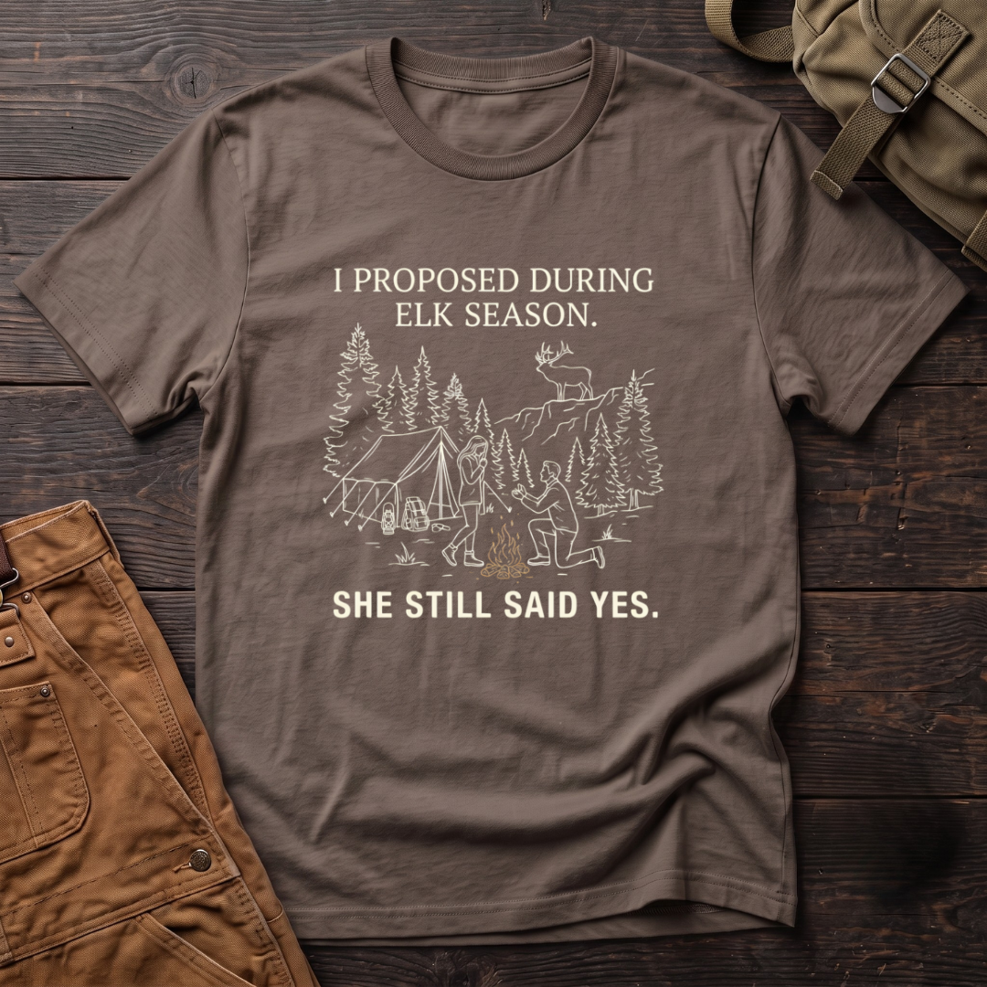 Brown funny elk hunting t-shirt with I Proposed During Elk Season, She Still Said Yes text and a line-art hunter kneeling by the campfire at an elk camp tent, with a bull elk on the ridge