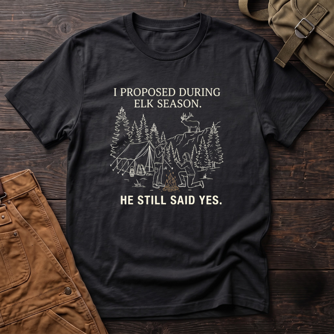 Black funny elk hunting t-shirt with I Proposed During Elk Season, He Still Said Yes text and a line-art hunter kneeling by the campfire at an elk camp tent, with a bull elk on the ridge