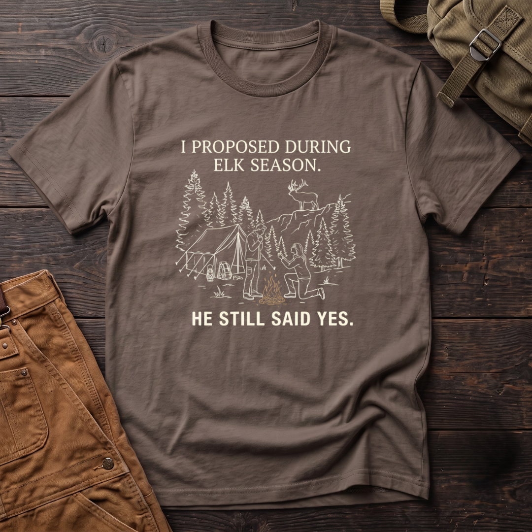 Brown funny elk hunting t-shirt with I Proposed During Elk Season, He Still Said Yes text and a line-art hunter kneeling by the campfire at an elk camp tent, with a bull elk on the ridge