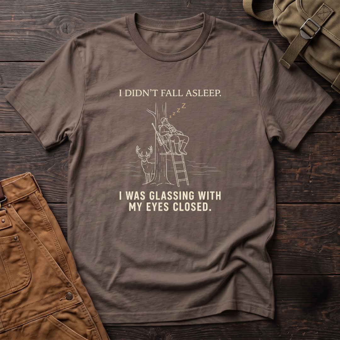 Brown funny deer hunting t-shirt with I Didn't Fall Asleep, I Was Glassing With My Eyes Closed text and a line-art hunter dozing in a ladder stand while a whitetail buck stands below