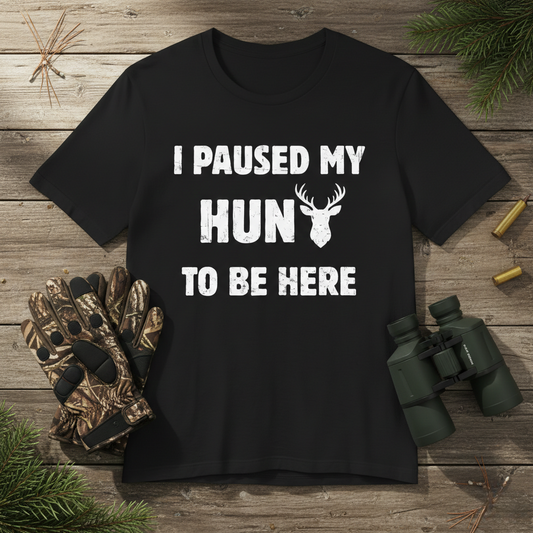 Funny deer hunting humor t-shirt in black laid flat on a wood surface beside camo gloves, binoculars, and spent bullet casings with pine sprigs.
