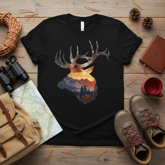 Bull elk mountain sunset double exposure graphic t-shirt in black laid flat on a wood surface beside binoculars, a canvas pack, hiking boots, a map, and a coffee mug.